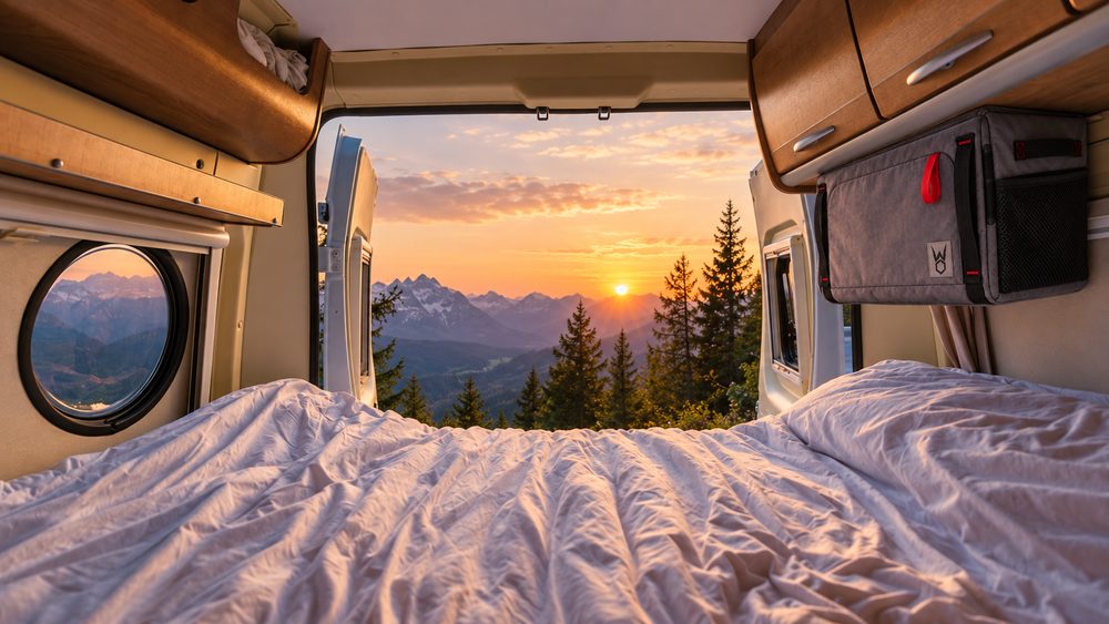 KI-Visualisierung: Blick aus einem Campervan auf Berge bei Sonnenuntergang, rechts hängt das Faultier unter dem Oberschrank.