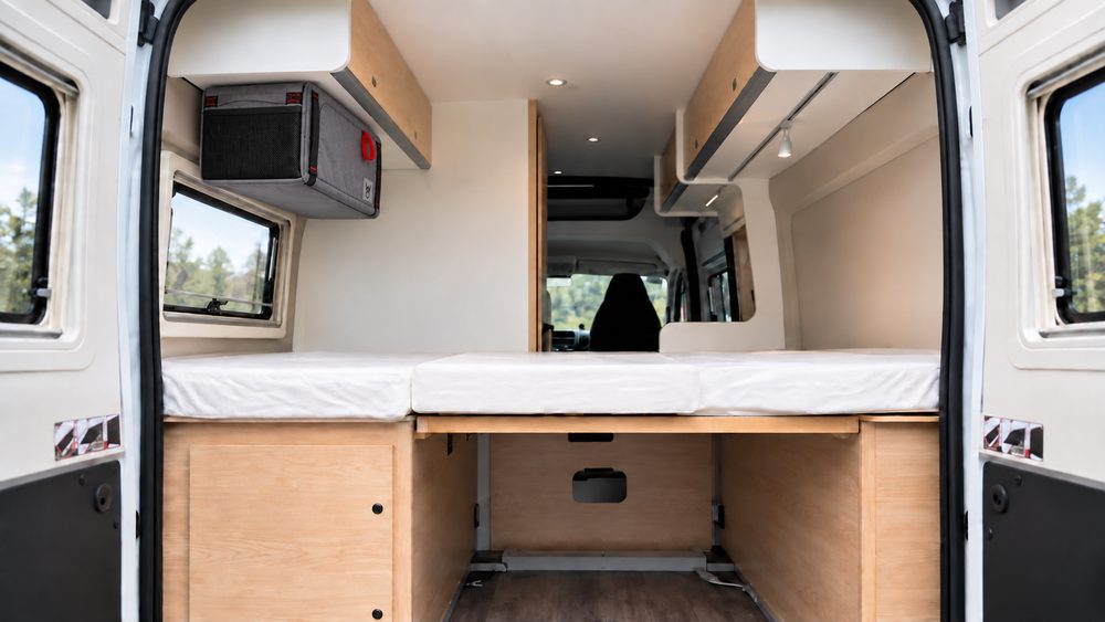 KI-Visualisierung: Das Faultier hängt unter einem Oberschrank im Heck eines hellen Campervans über dem Bett.